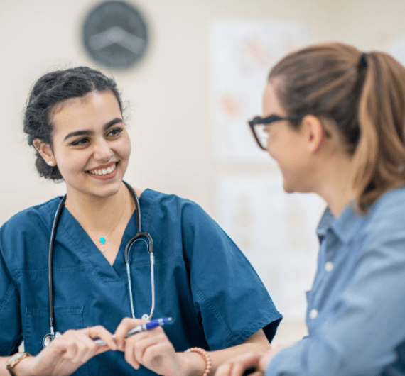 A 23 year old middle eastern woman with brown hair and a blue uniform talking to a 30 year old caucasian woman with glasses in a doctor’s office Why Choose HealthbridgeMD for Pharmacy Billing?
