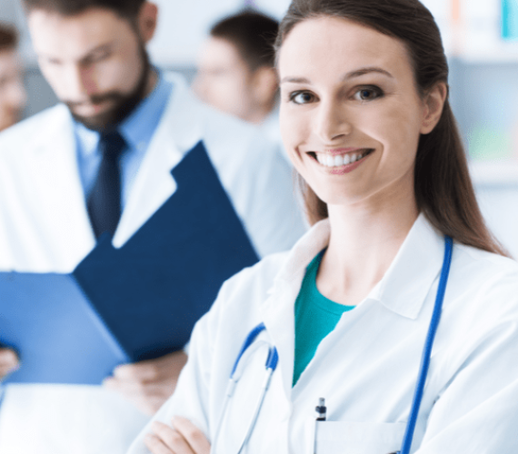 A caucasian woman , 30 years old, white hair, wearing a white coat and stethoscope, smiling, in a hospital with other doctors in the background Comprehensive Medical Credentialing Services