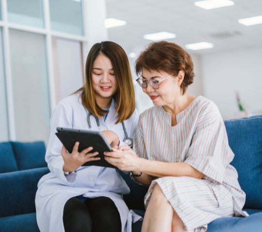 An asian woman doctor, 25 years old, showing a tablet to an asian woman patient, 70 years old, sitting on a blue sofa Revenue Cycle Analytics and Reporting