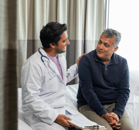 Two people, an indian man of 50 years old with a white coat and a clipboard, and an indian man of 30 years old with a blue sweater in a hospital room How Our Patient Benefits & Eligibility Verification Works